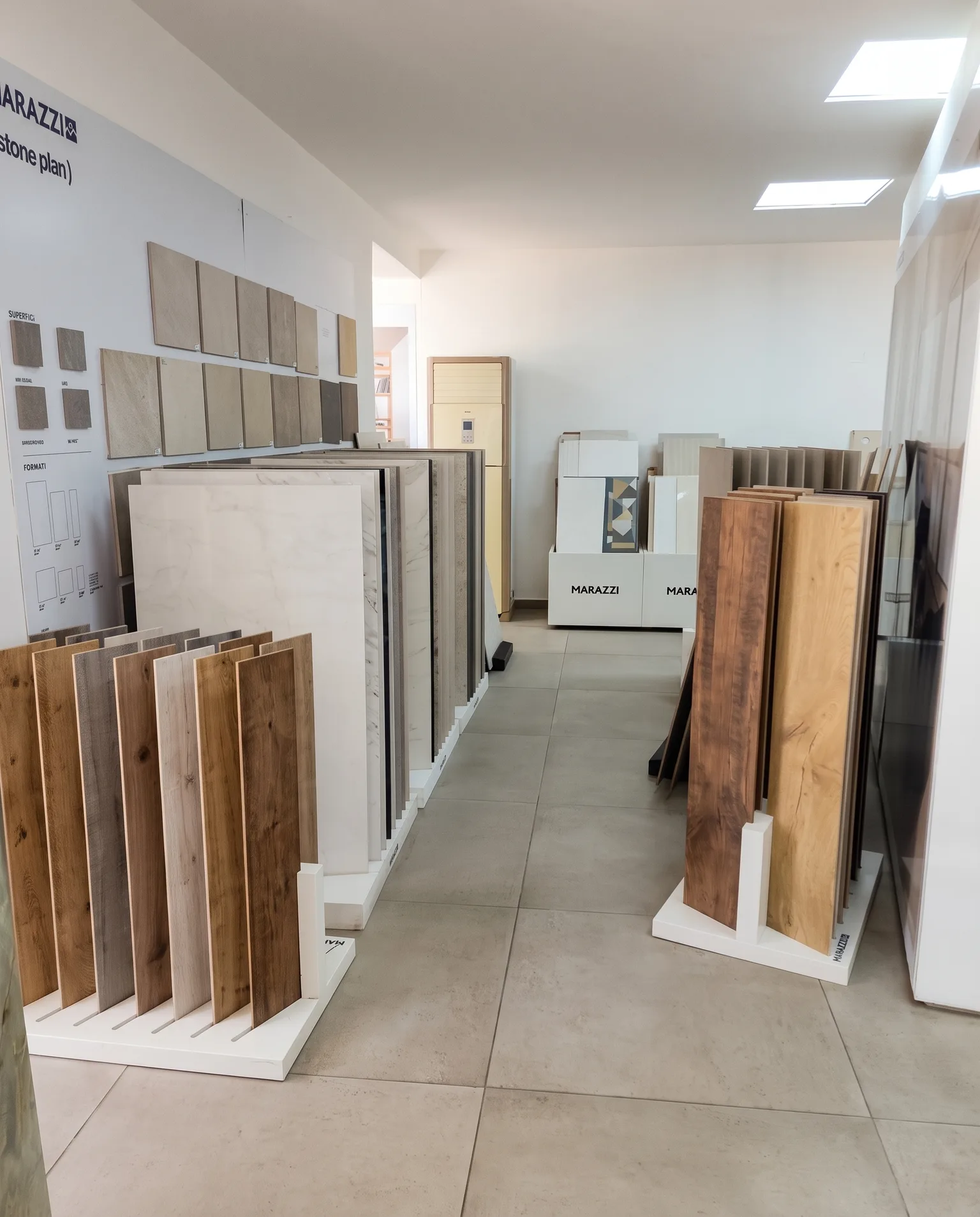 A showroom aisle lined with upright racks of stone- and wood-effect porcelain tiles on a pale tiled floor, Marazzi displays on the wall