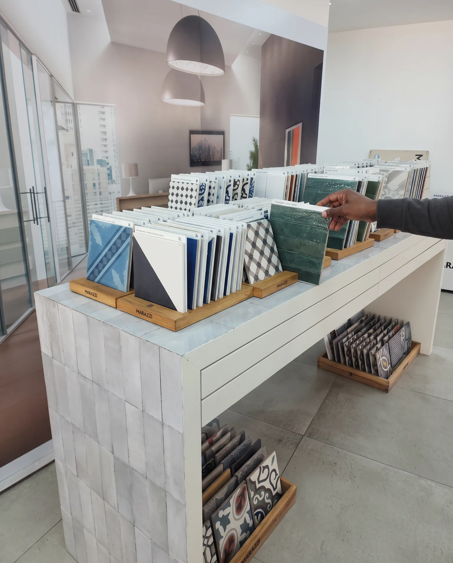 A hand lifting a patterned tile sample from a display table of arranged samples